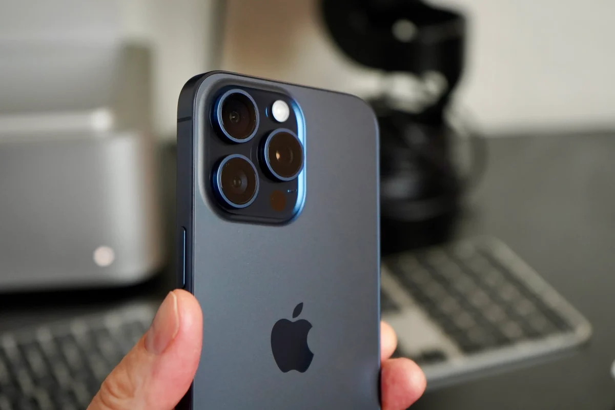 A person holds a smartphone with a blue finish, showcasing three camera lenses and an apple logo. In the background, a blurred keyboard and other electronics evoke the question: How to Fix Common iPhone Issues?.