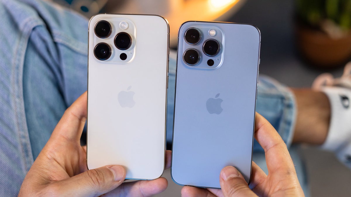 Two hands holding iPhone 13 vs iPhone 14 side by side. The left iPhone has a silver finish, while the right one is light blue. Both boast triple rear cameras. A person wearing jeans and white sneakers stands visible in the blurred background.