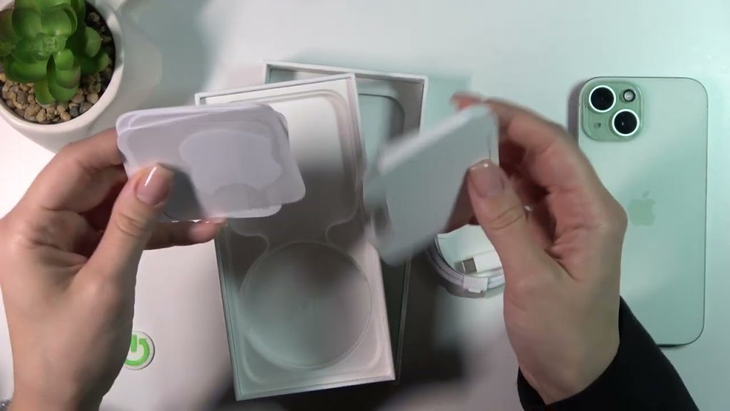 A person holds parts of an Apple product packaging, revealing included accessories and a phone, with a small plant in the background.