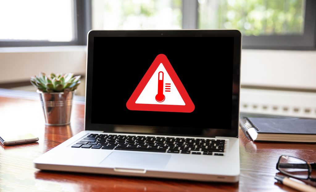 A laptop on a wooden desk displays a red alert symbol of a thermometer, indicating overheating. Green plant and stationery nearby.