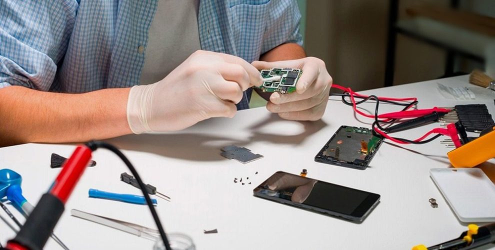 A technician in gloves repairs a smartphone circuit board, surrounded by tools and components on a cluttered workbench.