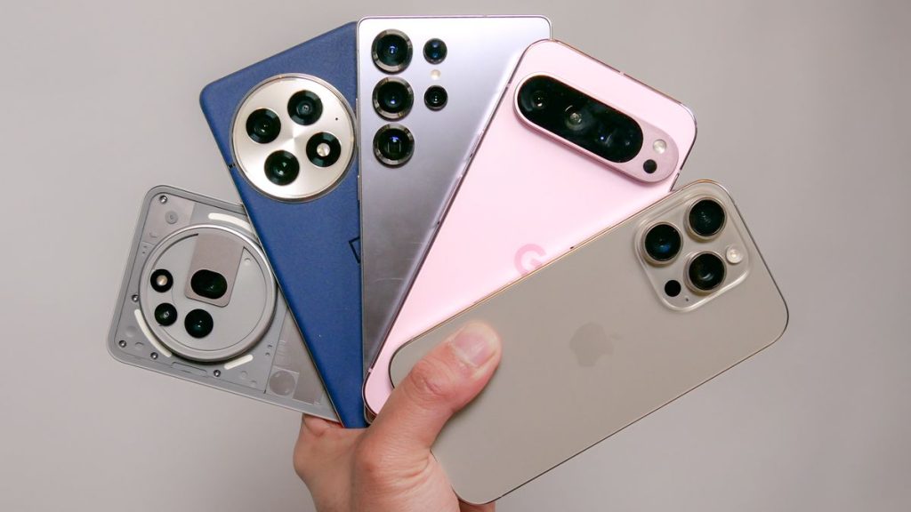 A hand holding five smartphones of various colors and designs, showcasing their distinct camera arrangements and features.