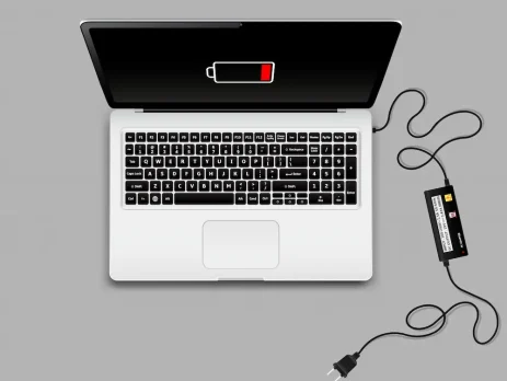 A laptop with a low battery warning on the screen, plugged into a power adapter on a gray background.