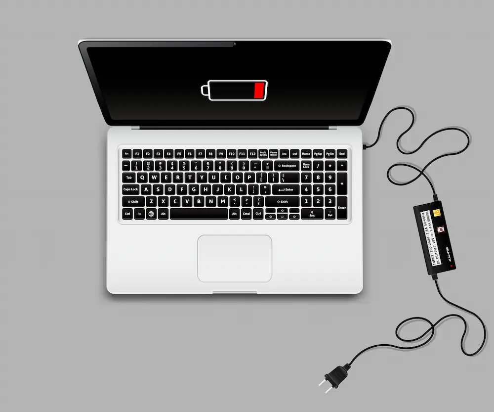 A laptop with a low battery warning on the screen, plugged into a power adapter on a gray background.