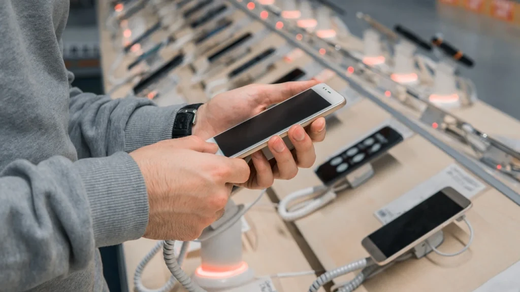 A person inspects a smartphone in a tech store, surrounded by other phones on display, with lighting illuminating their features.