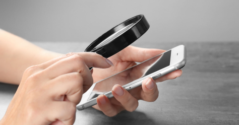 A hand holding a smartphone, while a magnifying glass hovers above the screen, highlighting a focus on detail.