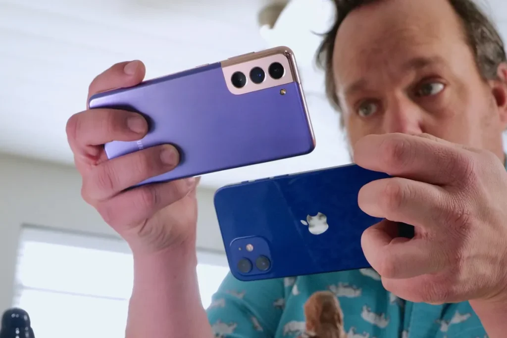 A person holding a purple Samsung smartphone and a blue iPhone, closely comparing their camera performance. The scene is set indoors with natural light streaming through a window in the background.