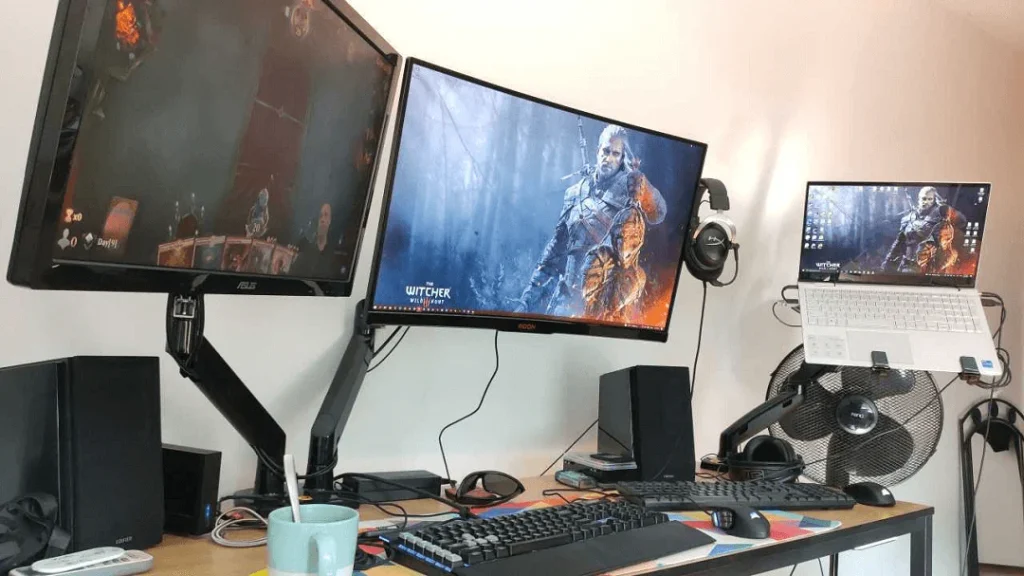 Three monitors on a desk: two large screens—perfect for Comparing Curved and Flat Monitors—display The Witcher wallpaper, while a laptop sits elevated on a stand. The setup includes speakers, keyboard, mouse, headphones, cup, and fan.