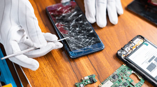A person wearing white gloves repairs a smartphone with a cracked screen using tools on a wooden table, offering comprehensive repair services surrounded by phone components and circuit boards.