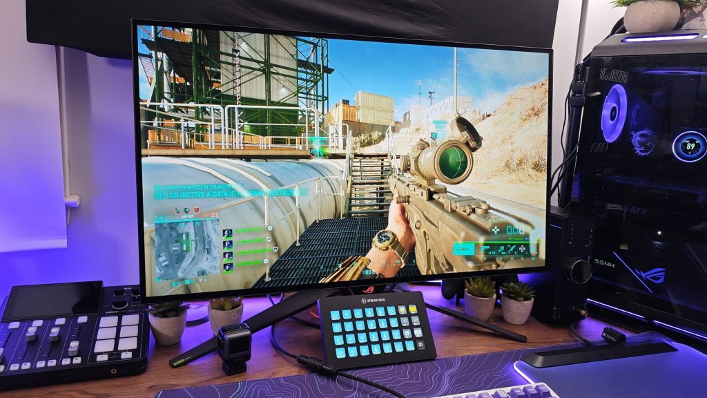 A computer monitor displays a first-person shooter video game. The desk features a stream deck, keyboard, watch, and audio mixer to help improve your gaming experience, with potted plants and an RGB-lit gaming PC in the background