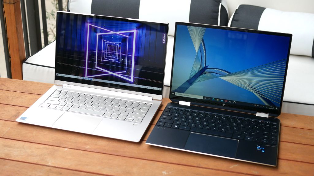 Two slim, modern laptops sit side by side on a wooden table. Their screens show geometric backgrounds, while their silver and black finishes catch the eye. Enjoy an enhanced keyboard and trackpad experience as you work comfortably in a cozy setting.