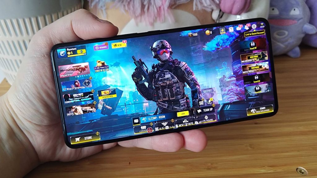 A hand holding a smartphone displaying a gaming interface with a soldier character, featuring various game modes and events.