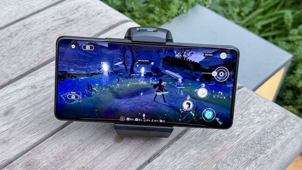 A mobile phone displays an animated video game scene with characters in a mystical landscape, resting on a wooden surface outdoors.