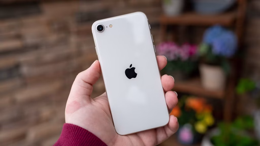 A hand holds a white iPhone SE (3rd Gen) with the Apple logo visible on the back. The background is slightly blurred, featuring shelves adorned with potted plants and flowers.